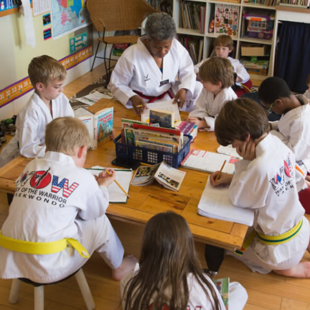 children dressed in taekwondo dobaks surrounding a table doing school work
